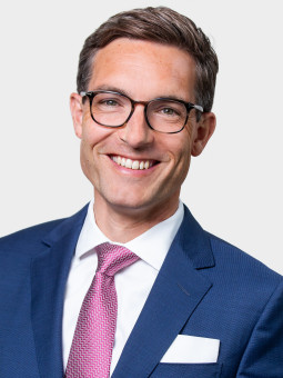 Mann mit kurzen, gewellten Haaren und Brille in dunkelblauem Anzug, weißem Hemd und rosa Krawatte, lächelt.