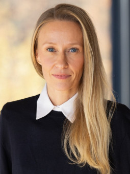 Porträt einer Frau mit langen, gewellten blonden Haaren, schwarzem Oberteil und neutralem Gesichtsausdruck.