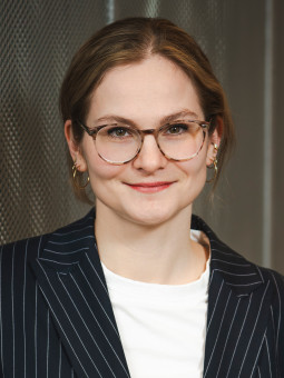 Mitarbeiterfoto: Friederike Micheael, Person mit kurzen, hellen Haaren, gestreiftem Blazer und Brille vor metallischer Wand.