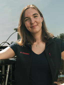 Person mit schwarzem Oberteil und Weste steht neben einem landwirtschaftlichen Gerät in ländlicher Umgebung.