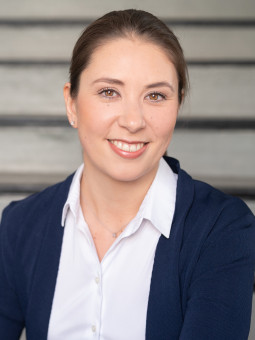 Frau mit Zopf sitzt auf einer Treppe, trägt weiße Bluse und dunkelblauen Blazer, lächelt in die Kamera.