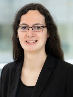 Person mit langen, dunklen Haaren in einem hellen Raum, trägt schwarzen Blazer und Brille, lächelt leicht.