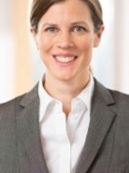 Frau mit braunem Pferdeschwanz, grauem Anzug und weißem Hemd, lächelt direkt in die Kamera. Heller, unscharfer Hintergrund.