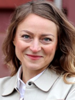 Frau mit langen, welligen, blonden Haaren, beigem Mantel und weißem Hemd, lächelt vor unscharfem roten Hintergrund.