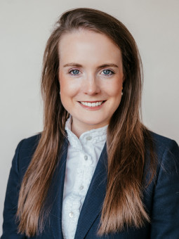 Carolin Weyers, Frau mit langen braunen Haaren, blauer Blazer, weißer Bluse, lächelt vor hellem Hintergrund.