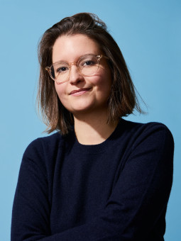 Person mit schulterlangen, braunen, gewellten Haaren, in dunkelblauem Pullover, vor hellblauem Hintergrund.