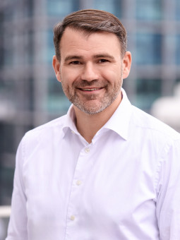 Dr. Christian Weber in a white shirt, smiling in front of modern buildings with large windows. Urban, bright setting.