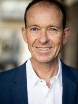 Mann mit kurzen, braunen Haaren, hellem Hemd und dunklem Blazer, lächelt in unscharfer städtischer Umgebung.