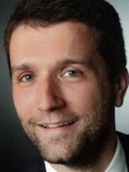 Mann mit kurzen, dunklen Haaren und Bart, trägt dunklen Anzug und helles Hemd, lächelt in die Kamera.