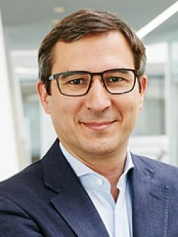 Mann mit Brille, dunklem Haar, in dunkelblauem Blazer und hellblauem Hemd vor moderner Architektur mit großen Fenstern.