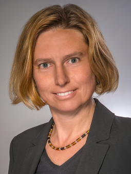 Person mit schulterlangen blonden Haaren, schwarzem Blazer und bunter Halskette, lächelt in die Kamera.