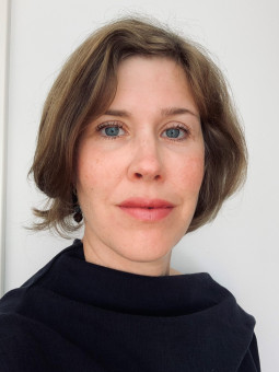 Frau mit kurzen, gewellten braunen Haaren, blauen Augen und schwarzem Oberteil vor hellem, einfarbigem Hintergrund.