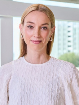 Lea Siering steht vor großen Fenstern, trägt ein weißes Oberteil und grüne Hose. Bäume und Gebäude sind im Hintergrund sichtbar.