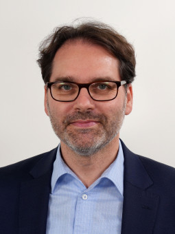 Mann mit dunklen, gewellten Haaren und grauem Bart in dunkelblauem Anzug und hellblauem Hemd, neutraler Gesichtsausdruck.