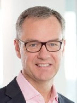 Mann mit Brille, grauen Haaren, dunklem Sakko und rosa Hemd, lächelt in die Kamera vor unscharfem Hintergrund.