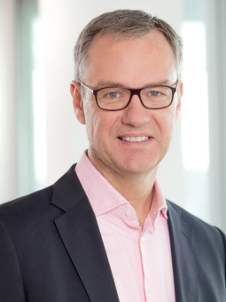 Mann mit Brille, grauen Haaren, dunklem Sakko und rosa Hemd, lächelt in die Kamera vor unscharfem Hintergrund.