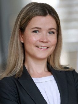 Frau mit langen blonden Haaren in schwarzem Blazer und weißem Oberteil, lächelt an einer Wand stehend.