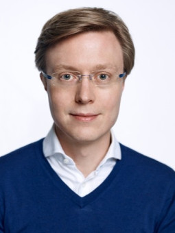 Mann mit kurzen, hellbraunen Haaren, blauer Pulloverjacke und schmaler Brille, blickt direkt in die Kamera.