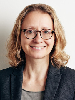 Frau mit lockigem, blondem Haar, grauer Bluse und dunklem Blazer, lächelt in die Kamera. Heller, einfarbiger Hintergrund.