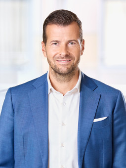 Mann in blauem Anzug und weißem Hemd steht in modernem Innenraum mit großen Fenstern. Hände in Hosentaschen.