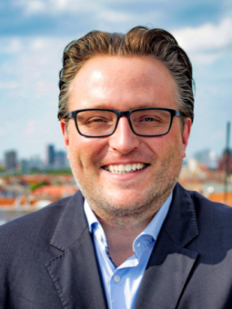 Mann mit kurzen, gewellten Haaren und Brille, trägt dunklen Blazer und hellblaues Hemd, lächelt in die Kamera.