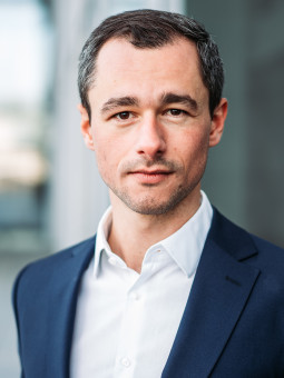 Mann mit dunklen Haaren und Bart in dunkelblauem Anzug und weißem Hemd, blickt in die Kamera. Unscharfer Hintergrund.