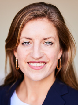 Frau mit langen, braunen Haaren, dunklem Blazer und auffälligen Ohrringen, lächelt vor unscharfem, buntem Hintergrund.