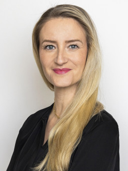 Verena Thaler, Frau mit langen blonden Haaren, schwarzem Oberteil, verschränkten Armen, vor weißem Hintergrund.