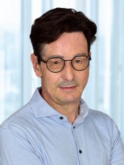 Mann mit dunklen, gewellten Haaren und Brille in hellblauem Hemd, Arme vor der Brust, helle Innenarchitektur.