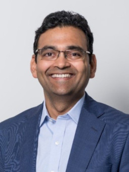 Mann mit dunklen Haaren und Brille in blauem Anzug und hellblauem Hemd, lächelt vor einfarbigem Hintergrund.