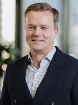 Mann mit kurzen, hellen Haaren in modernem Raum, dunkler Blazer, helles kariertes Hemd, lächelt in die Kamera.