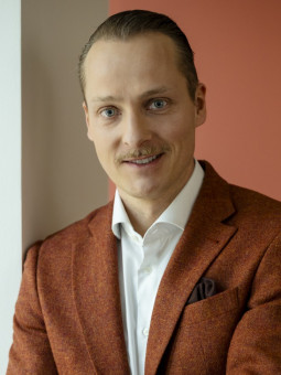 Mann mit kurzem Haarschnitt und Schnurrbart in braunem Blazer und weißem Hemd vor zweifarbiger Wand.