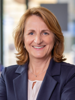 Frau mit schulterlangen, lockigen, blonden Haaren in einem hellen Raum, trägt dunkelblauen Blazer und lächelt.