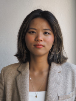 Junge Frau mit schulterlangen, gewellten Haaren in Blazer, blickt neutral in die Kamera vor heller Wand.