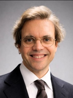 Mann mit hellen, lockigen Haaren, Brille, dunklem Anzug und Krawatte lächelt vor hellem Hintergrund.