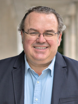 Mann in hellem Hemd und dunklem Blazer mit Brille, lächelnd, Hände in Hosentaschen, unscharfer grüner Hintergrund.