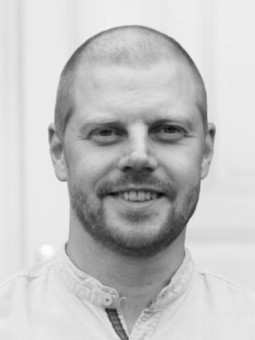 Mann mit kurzem Haar und Bart, trägt helles Hemd, lächelt freundlich in die Kamera. Hintergrund unscharf.