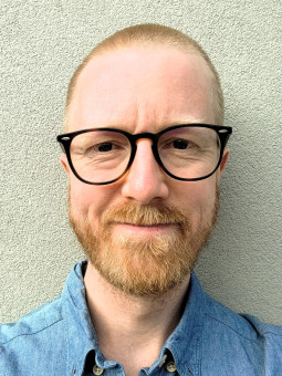 Mann mit kurzem, blonden Bart und Glatze, trägt blaue Hemdbluse und schwarze Brille, lächelt vor grauer Wand.