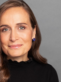 Frau mit langen, braunen Haaren, schwarzem Oberteil und blauem Ohrring vor grauem Hintergrund. Leichtes Lächeln.