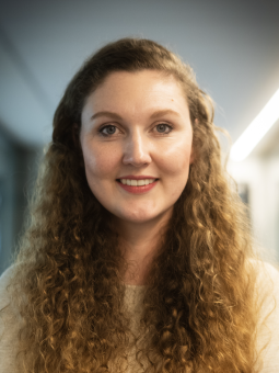 Frau mit langen, lockigen Haaren in hellem Pullover, lächelt mit verschränkten Armen, trägt eine goldene Armbanduhr.