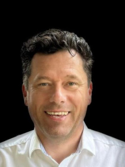 Mann mit dunklen, lockigen Haaren und freundlichem Lächeln, trägt helles Hemd, schwarzer Hintergrund.