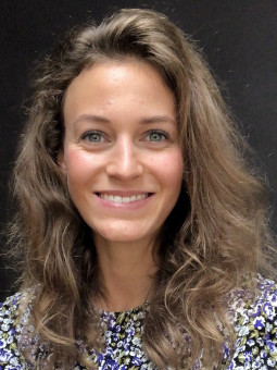 Frau mit langen, lockigen, braunen Haaren und buntem Blumenmusterkleid vor schwarzem Hintergrund.