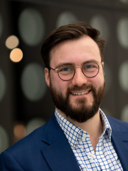 Mann mit Bart und Brille in blauem Sakko vor unscharfem, dunklem Hintergrund mit hellen Lichtpunkten.