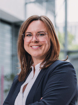 Frau mit langen, braunen Haaren in dunkler Jacke und Brille, lächelt in modernem Innenraum mit Tageslicht.