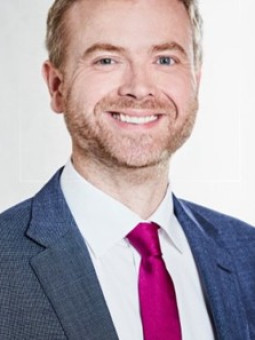 Mann mit blonden Haaren, Bart, dunkelblauem Anzug und pinker Krawatte lächelt in die Kamera. Heller, neutraler Hintergrund.