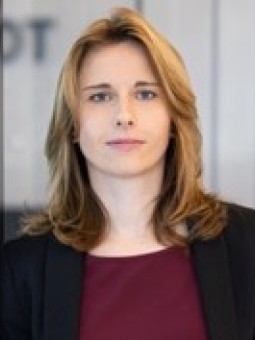 Person mit langen, blonden Haaren in schwarzem Blazer und burgunderfarbenem Oberteil vor transparenter Wand mit „COT“.