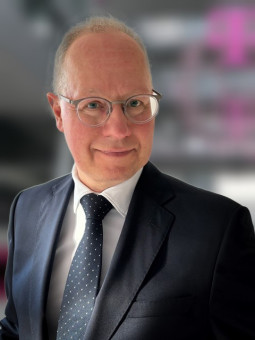 Mann mit Brille in formellem Anzug, lächelt, steht seitlich, unscharfer moderner Hintergrund.