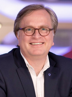 Mann mit Brille lächelt in dunklem Anzug und weißem Hemd vor unscharfem, hellem Hintergrund.