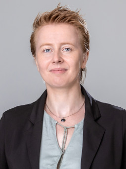 Anne-Marie Tumescheit in schwarzem Blazer und hellgrünem Oberteil, lächelnd vor grauem Hintergrund.
