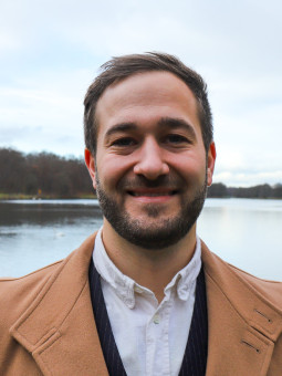 Mann mit kurzem Bart und beigem Mantel steht vor einem ruhigen See, umgeben von Bäumen und Steinen im Wasser.